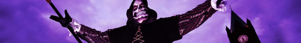 A person in dark, skeletal-themed costume raises their arms. They stand against a purple sky with a clock tower in the background.