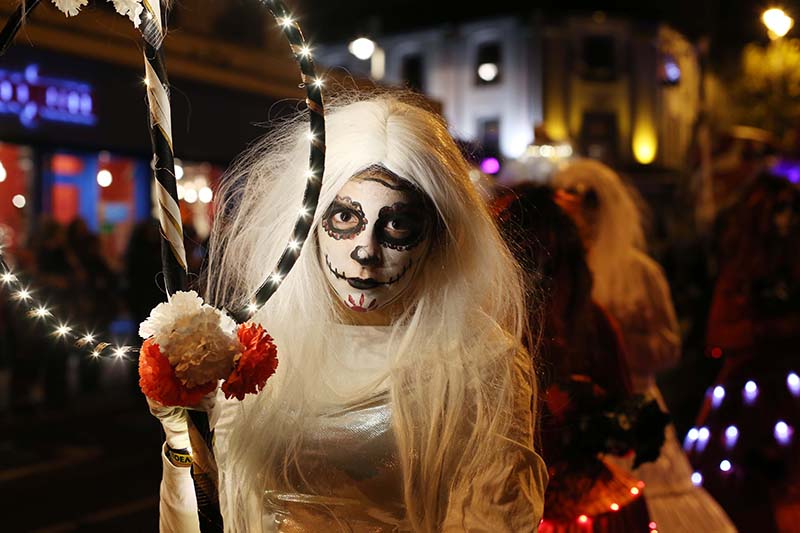 Person in white costume with skeleton face paint holds a hoop wrapped in lights and flowers at a nighttime event. Buildings and people are visible in the background.