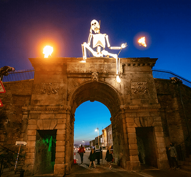 Stone archway with a large skeleton figure and flaming torches on top. People walk underneath. Twilight sky in the background.