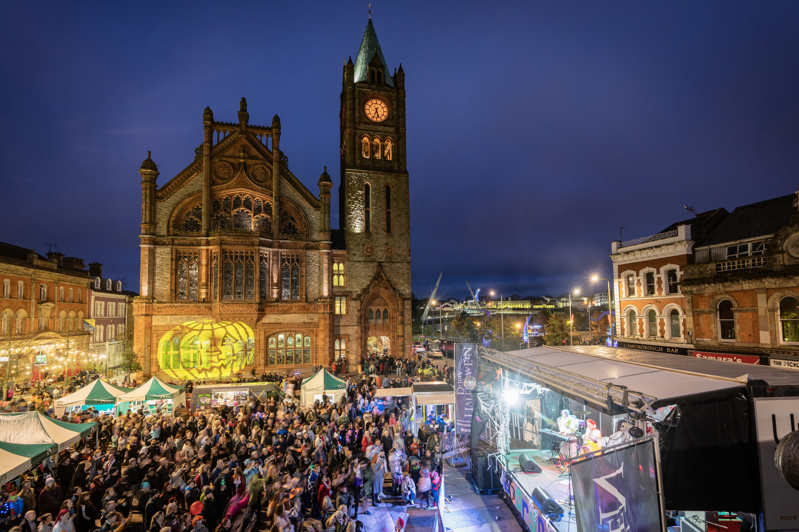 A large crowd gathers for family fun at an outdoor festival in front of a historic church with a clock tower at dusk, surrounded by market stalls and lit buildings.