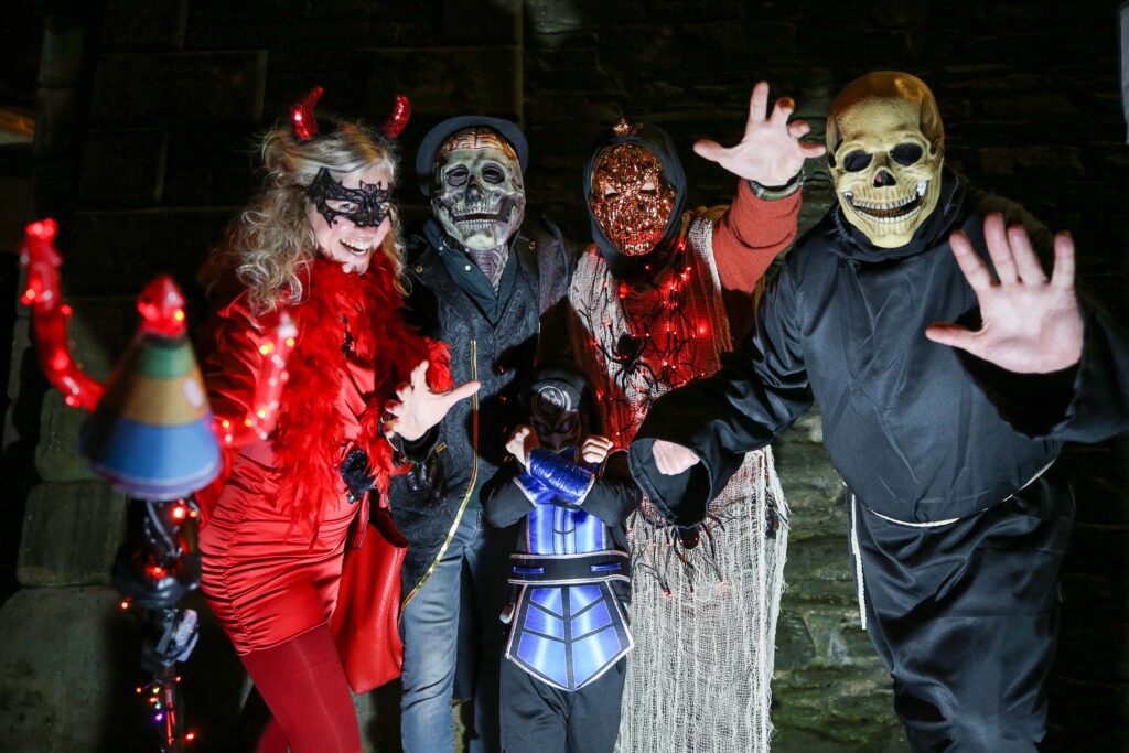 A group of five people in various affordable Halloween costumes, including devils, skeletons, and masks, pose together at night against a stone wall.
