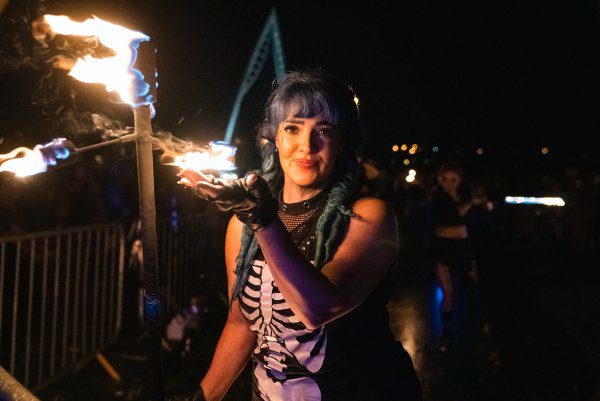 A woman with blue hair and a skeleton costume holds a flaming torch with her gloved hand at an outdoor night event.