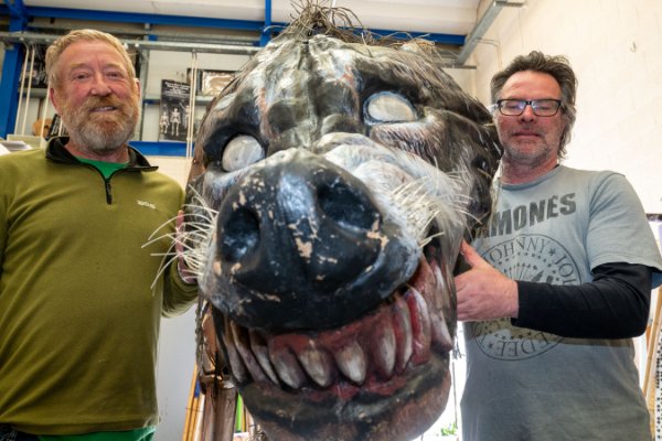 Two men stand indoors holding a large, detailed, and somewhat worn wolf head prop with exaggerated teeth and eyes.