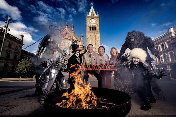 A group of people in Halloween costumes pose around a fire pit in a city square with historic buildings and a clock tower in the background.