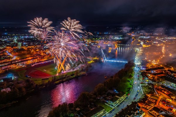 Aerial view of a city at night with fireworks lighting up the sky over a river, bridges, and illuminated buildings.