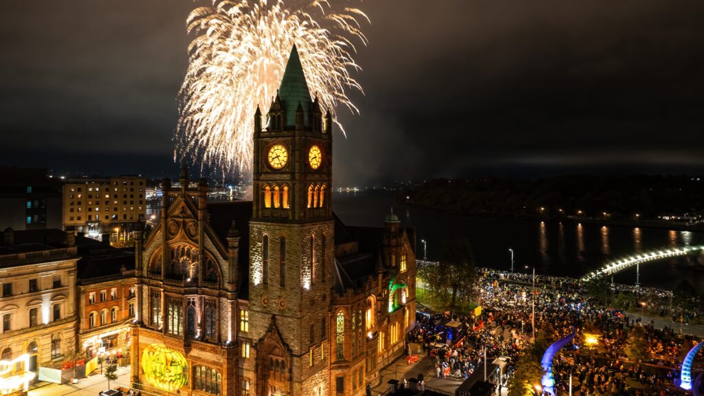 Fireworks light up the night sky above a large historic clock tower, with a crowd of people gathered around the building and nearby streets. Some in attendance whisper about the Ghost of the Guildhall, recalling tales like the Legend of Sam Mackey as they watch the colourful bursts illuminate both stone and story.